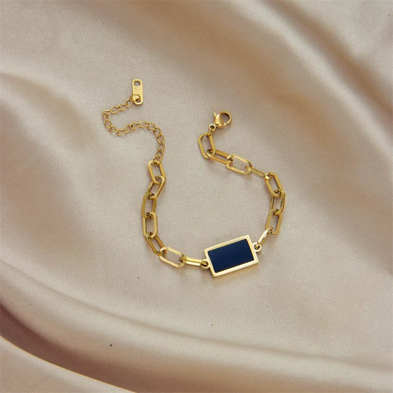 Gold chain bracelet with a blue square charm on a beige fabric background